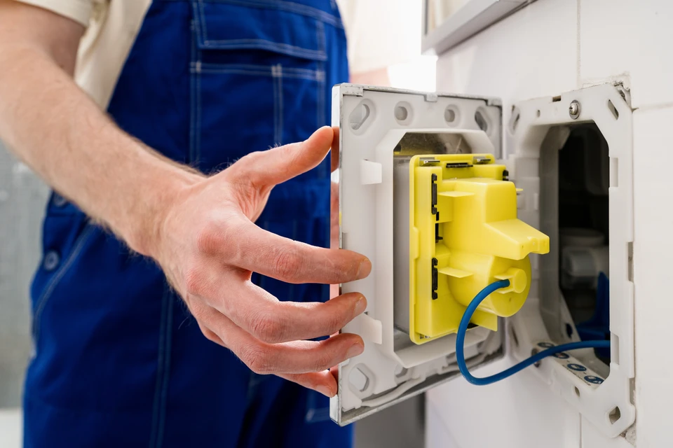 Un plombier en salopette bleue et chemise à carreaux répare un robinet de cuisine dans un intérieur moderne avec mur en briques blanches